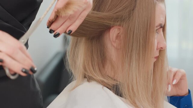 Blonde caucasian woman getting haircut, stylist sectioning and combing hair with black nails, closeup of scissors trimming blunt ends in bright modern salon, focused calm atmosphere and careful