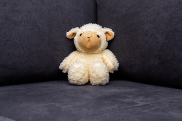 Fototapeta premium Cute plush lamb toy sitting on a dark sofa. Minimal cozy interior scene, childhood, comfort and nursery decor concept with copy space.