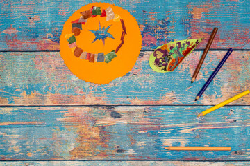 Top view of handmade paper sun and leaf crafts with pencils on a blue wooden background.