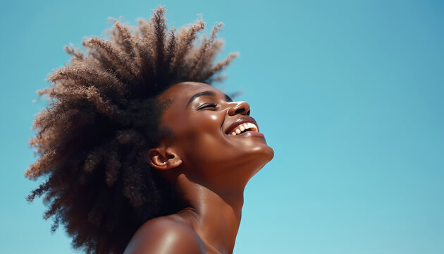 Black woman smiles with eyes closed outdoors against a clear blue sky. Her head is tilted back in a moment of pure joy and peace, hair blowing in wind. Freedom and happiness.