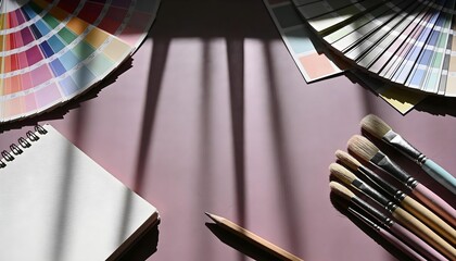 Dramatic artist workspace with a fan of color swatches, paintbrushes, and a notebook under high-contrast window shadows.