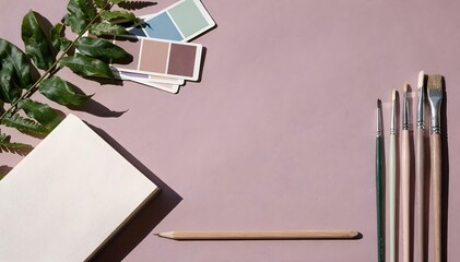 Top view of an art studio desk with paintbrushes, an open notebook, and botanical leaves on a dusty rose surface.