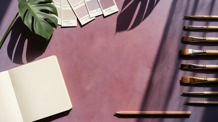 Creative art studio workspace with paintbrushes, a blank notebook, color swatches, and a monstera leaf on a plum background.