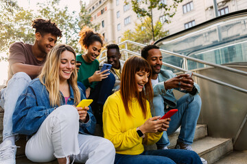 Multiracial group of millennial student friends sitting together outdoors, watching social media content on a smart phone and laughing joyfully. Youth and technology lifestyle concept.