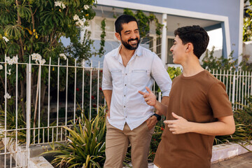 Father and teenage son smile while talking during a walk in a small residential neighborhood,...