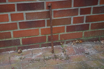 Rusty metal stake leaning against a brick wall above a paved walkway with weeds and dry leaves. An old outdoor detail with decay.