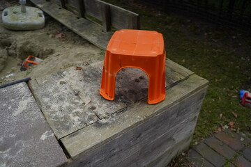 Orange plastic stool on a worn wooden sandbox edge beside sand and yard pavers. A small outdoor...