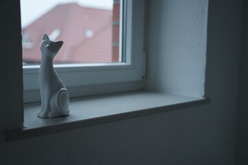Small ceramic cat figurine on a windowsill beside a curtain, with a blurred roof outside the window. Quiet indoor still life scene.