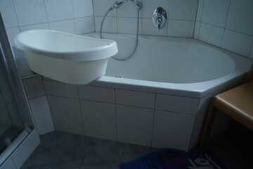 White bathtub with a curved wall-mounted sink and metal faucet in a tiled bathroom, with a shower panel on the left. Simple bathroom fixtures in a clean setting.