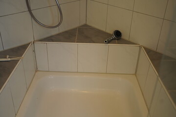 White bathtub with tiled walls, corner ledge, shower hose, and a metal pipe fixture. Clean bathroom interior with neutral tiles.