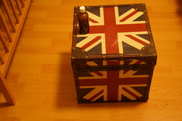 Wooden floor with a vintage trunk decorated in a red, white, and blue flag pattern, topped with a small brown bottle. A patriotic storage box sits indoors.