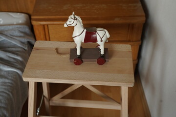 Wooden toy horse with red saddle on a small table beside a bed and dresser. Simple indoor still life scene.
