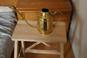 Brass watering can with long spout on a small wooden table beside a bed in a cozy room. Simple indoor still life with warm tones.