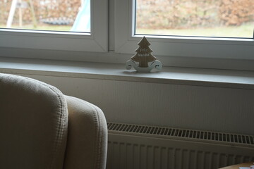 Interior window sill with a small tree-shaped decoration and "Noel" text, sofa edge and heater below; outdoor view blurred. Cozy home scene.