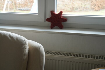 Red starfish-shaped plush toy on a windowsill above a radiator, with a sofa armrest in the foreground and an outdoor view through the window. Cozy indoor corner scene.