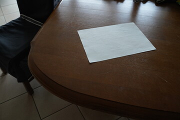 White paper sheet on a dark wooden table beside a padded chair in a tiled room. The scene shows a blank paper on a table.