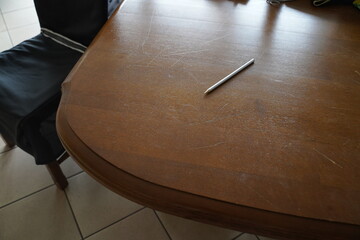 Scratched wooden table with a silver pencil lying on it beside a dark chair on tiled floor in daylight. An empty tabletop scene.