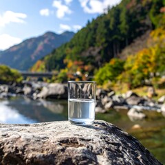 A glass of water sits on a rock by a river with trees and a bridge in the background on a sunny day