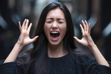 Portrait of a young brunette woman screaming in frustration or despair, hands raised, eyes closed, against a dark, blurred urban outdoor background
