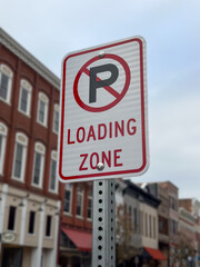 A loading zone sign with a no parking symbol. The background consists of blurry downtown brick buildings.