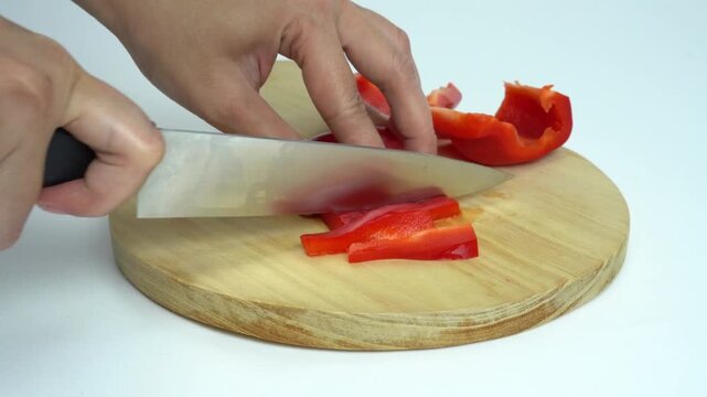 A woman is cutting the body of a red bell pepper or red pepper lengthwise like a matchstick, on a round brown wooden cutting board, isolated on a white background.