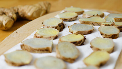 Ginger root slices air drying on white paper towel, whole root nearby on wooden cutting board, prepared for cooking or herbal remedy use