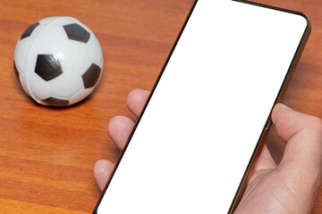 Close-up of a hand holding a cell phone with a blank screen next to a soccer ball as a concept for a sports app and sports betting