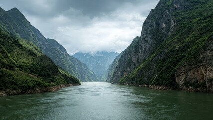 Fototapeta premium Deep river flowing between steep green mist shrouded mountains under a cloudy sky