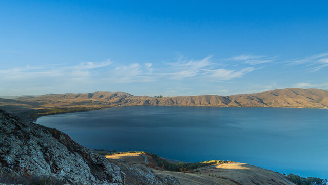 Tranquil landscape of Lake Sevan, Armenia seen from a rocky hillside. The deep blue water reflects the sky, surrounded by distant brown mountains.