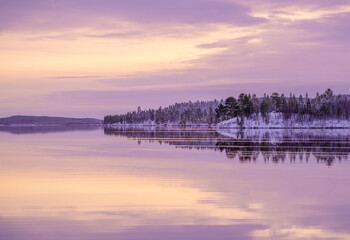 A winter scene shows a calm water surface reflecting a snow-covered island and trees. The sky is filled with purple hues at sunset, creating a quiet atmosphere.