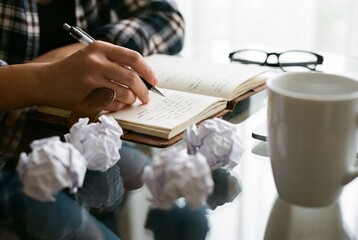 Woman hand writing with pen in open notebook near crumpled paper balls and white coffee mug on desk