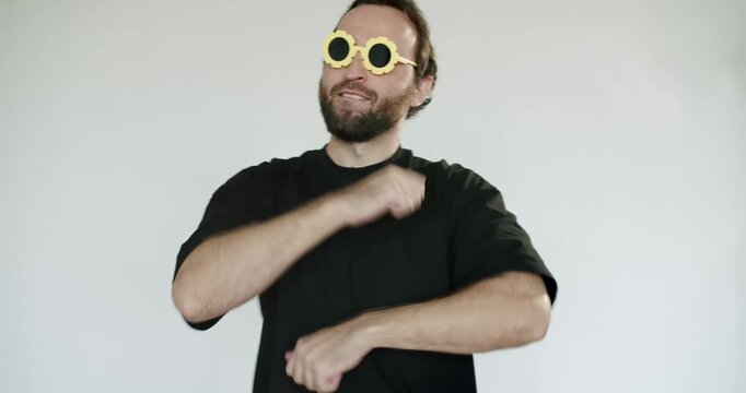 A man with a beard dances and celebrates while wearing a festive party hat and quirky sunglasses in a studio setting with simple background, expressing joy and excitement.birthday party or celebration