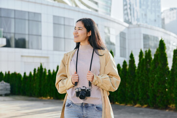 Woman with camera smiling outdoors in an urban setting, casual fashion, bright daylight, street...