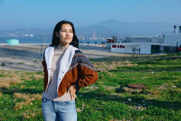 Woman standing in a coastal field wearing a patterned shawl over a knit sweater and jeans, sunny day, breezy atmosphere, distant hills and sea behind, confident pose.