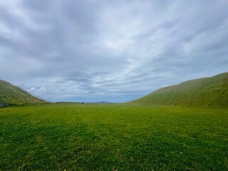 A panoramic view of a green dormant volcano crater and grass field under blue semi-cloudy sky.