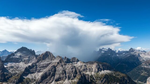 Majestic white clouds drift over snow-capped mountain peaks and rocky alps in this stunning summer landscape view under a clear blue sky