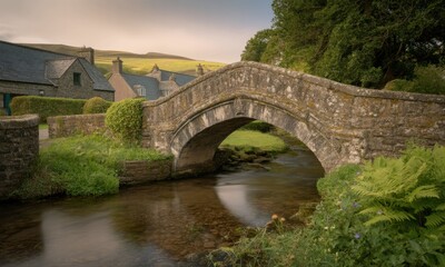 Fototapeta premium A quaint stone arch bridge spans a calm stream, nestled beside charming stone houses and lush greenery under a soft, dusky sky. Rolling hills form a picturesque backdrop
