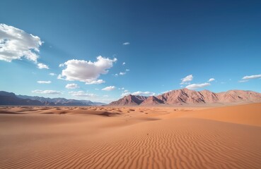Naklejka premium Vast desert landscape features orange sand dunes with wind ripples under bright blue sky with scattered clouds. Jagged mountains rise in distance across dry arid terrain. Wide open uninhabited