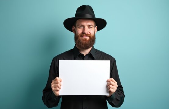 Bearded man in black shirt and hat holds white blank paper sheet. Simple background with copy space for text. Person displays information for ads or signs.