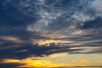 A dramatic evening sky is covered with thick blue and gray clouds, through which bright golden light from the setting sun breaks on the horizon.