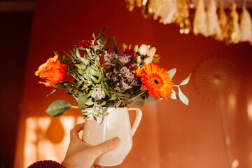 Hand holding flower bouquet in white jug against warm wall