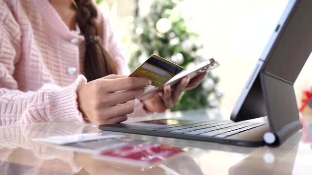 An Asian woman using a tablet while holding credit cards at a table indoors, representing online shopping, digital payment, and modern lifestyle in a casual home setting.