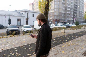 A man is using his phone while walking along a sidewalk covered with yellow leaves