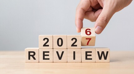 A hand carefully places a wooden block with the number seven onto others spelling out 2026 review signifying a transition