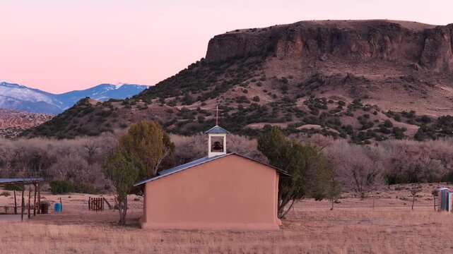 A cinematic drone orbit at a medium distance features a rural church cross and steeple at dusk. The shot circles to reveal a massive mesa and snowy peaks. Perfect for documentary opening sequences.