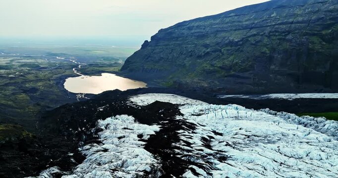 Drone flyover of an ash-streaked Iceland glacier with a distant mountain lake and steep cliff, showing melt patterns, moraine lines, and rugged volcanic terrain.