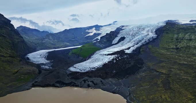 Wide drone view of Icelandic icecap spilling through a valley into a tan meltwater lagoon, with green slopes, dark lava gravel, and braided channels under overcast sky.