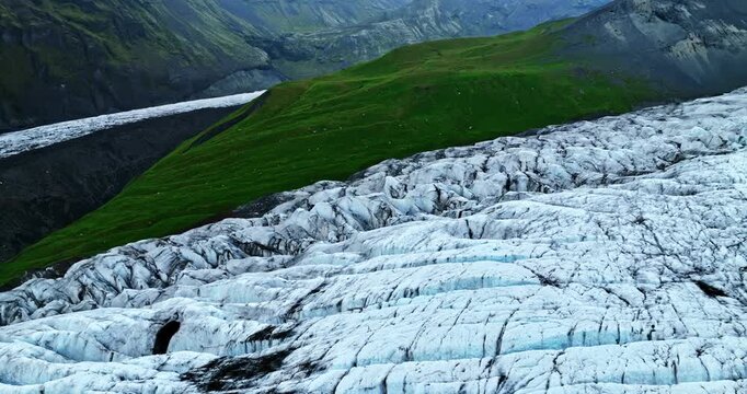 Aerial drone sweep over a rugged Iceland glacier, revealing deep crevasses and blue ice beside vivid green mountain slopes and a volcanic valley.