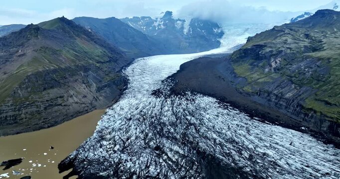 Drone aerial over an Iceland glacier tongue flowing through a volcanic valley toward a muddy meltwater lagoon, with green mountains and dark moraine ridges.