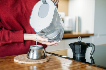 Young woman pours water from filter pitcher into base of moka pot on wooden kitchen island. Slow coffee movement, intentional brewing process, specialty coffee lifestyle.